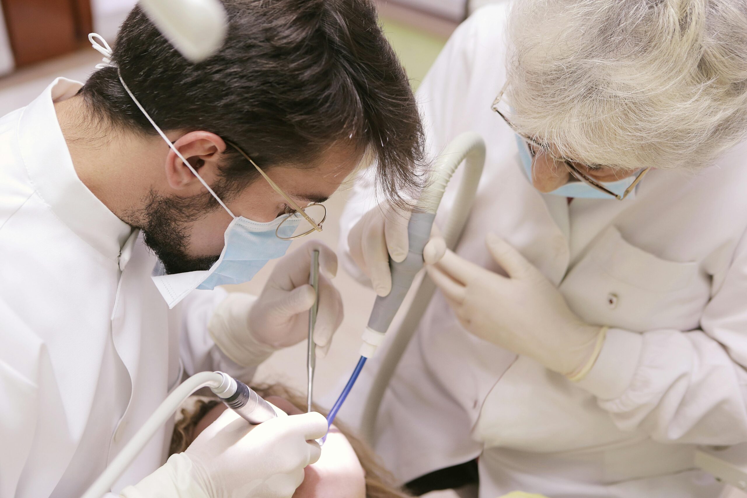 Two dental professionals wearing masks, gloves, and protective glasses work closely over a patient in a dental chair.