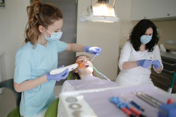 wo dental professionals wearing masks and gloves provide treatment to a patient reclining in a dental chair.