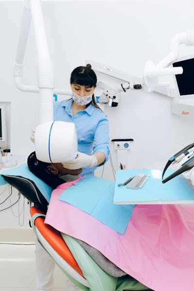 A female dentist in a blue uniform and floral face mask uses advanced medical equipment to examine a patient in a modern dental clinic.