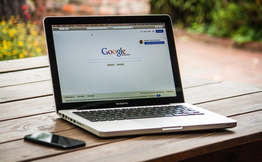 An open silver MacBook Pro laptop sitting on a wooden table outdoors, displaying the Google homepage on the screen. A black smartphone is next to it.