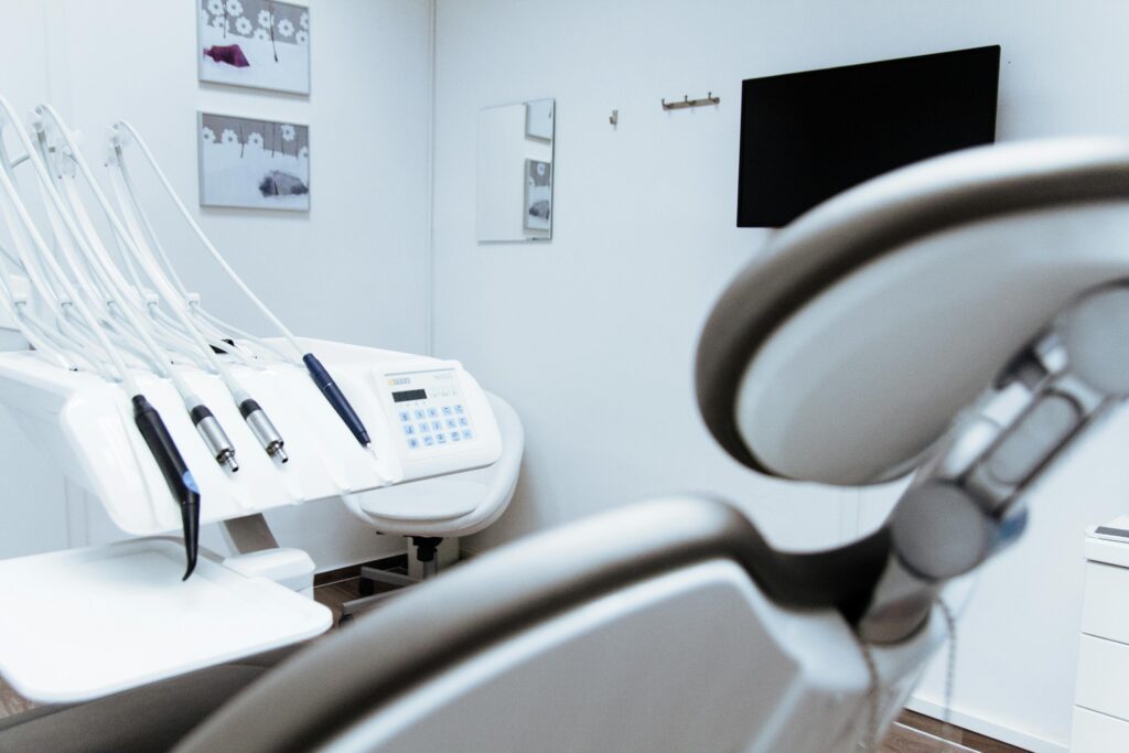 A clean, modern dental office operatory room with a white dental chair, various dental tools, a monitor on the wall, and pictures.