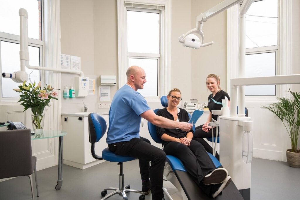 A male dentist speaks with a female patient in a modern dental office, assisted by a female dental assistant.