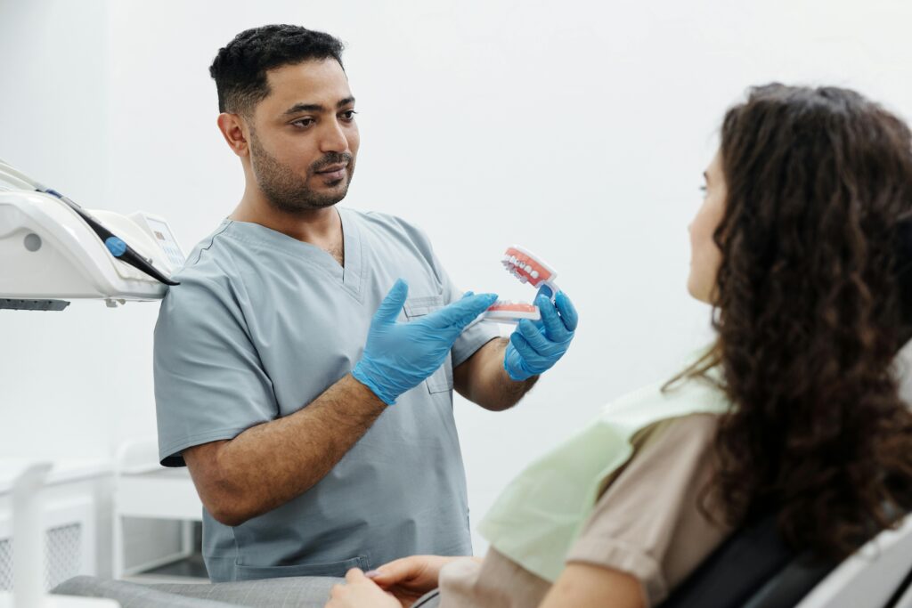 A male dental professional in gray scrubs and blue gloves holds a dental model of teeth and gums while explaining oral health to a female patient in a dental office.