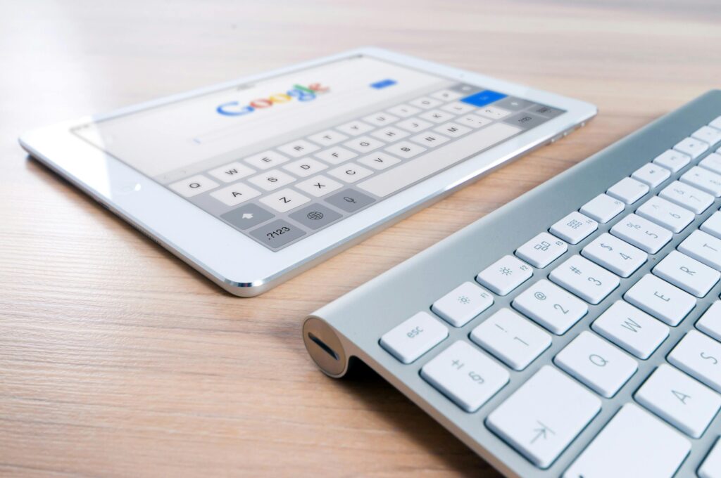 A white tablet displaying the Google search page and an on-screen keyboard sits next to a silver and white external physical keyboard on a light wooden desk.