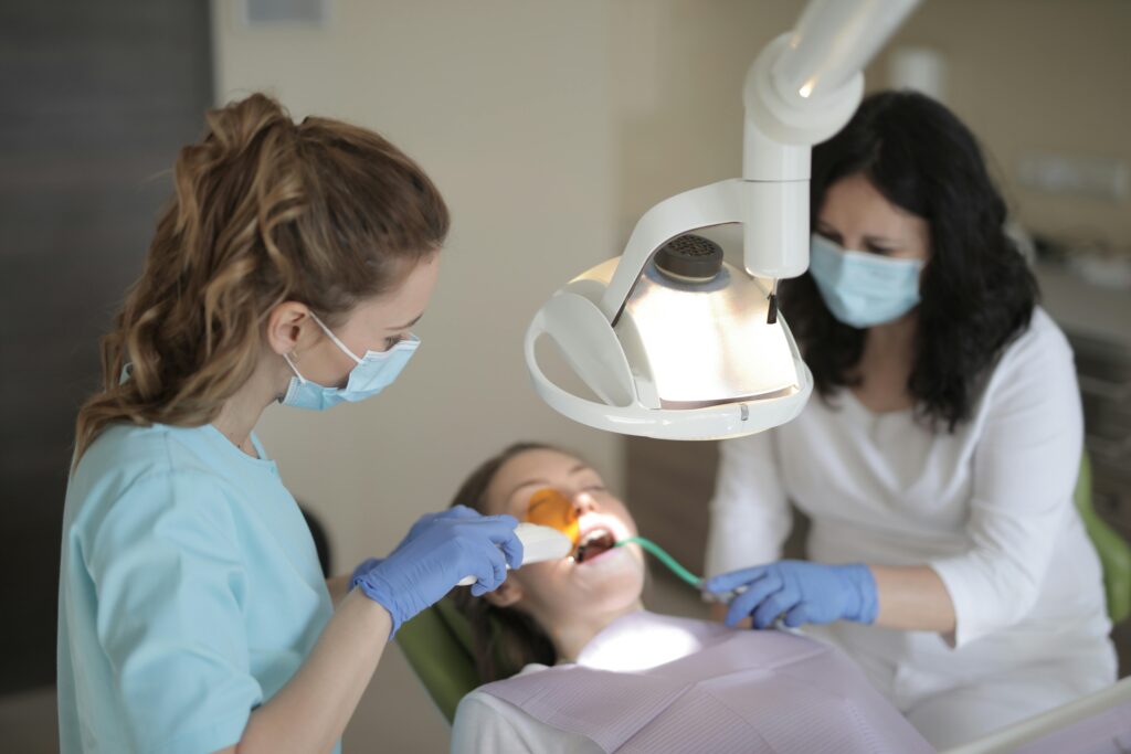Two dental professionals wearing masks and gloves perform a procedure on a patient in a dentist's chair, using a curing light.