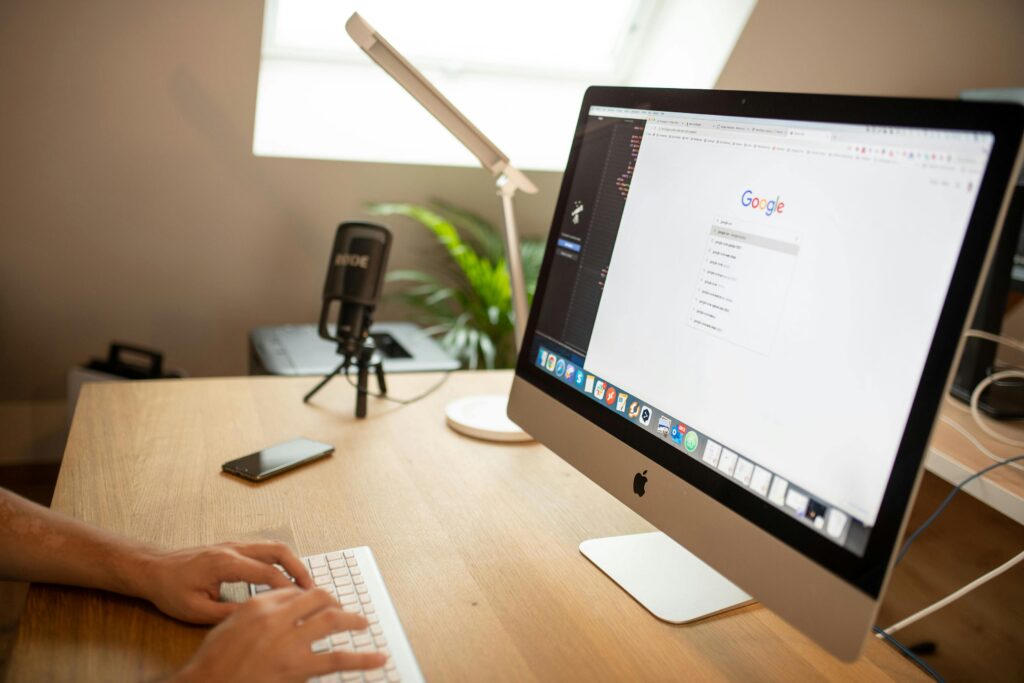A person typing on a white keyboard at a desk with an iMac computer showing the Google homepage, a microphone, and a smartphone.