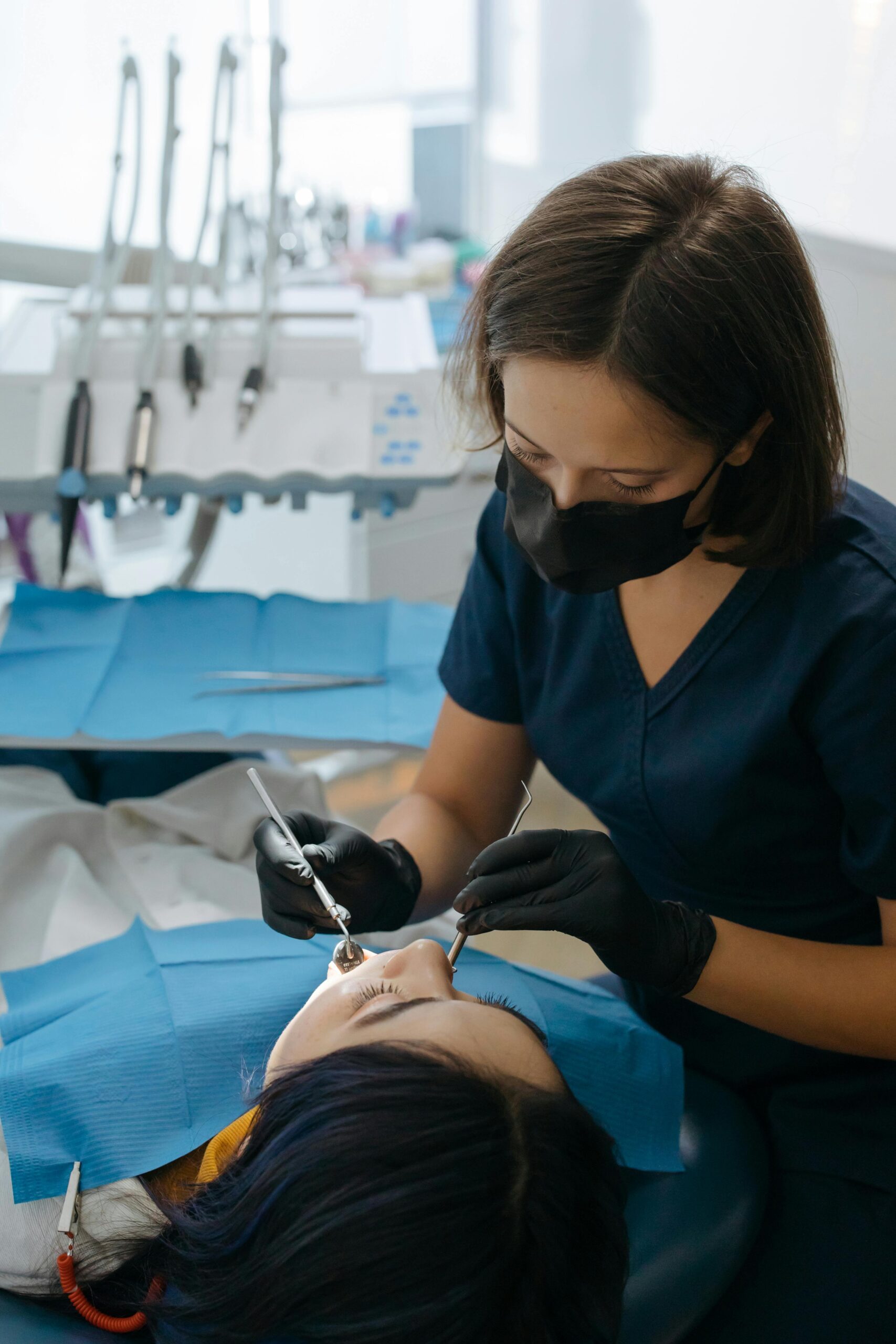 A female dentist wearing a face mask and black gloves performs a dental examination on a patient lying in a dental chair.